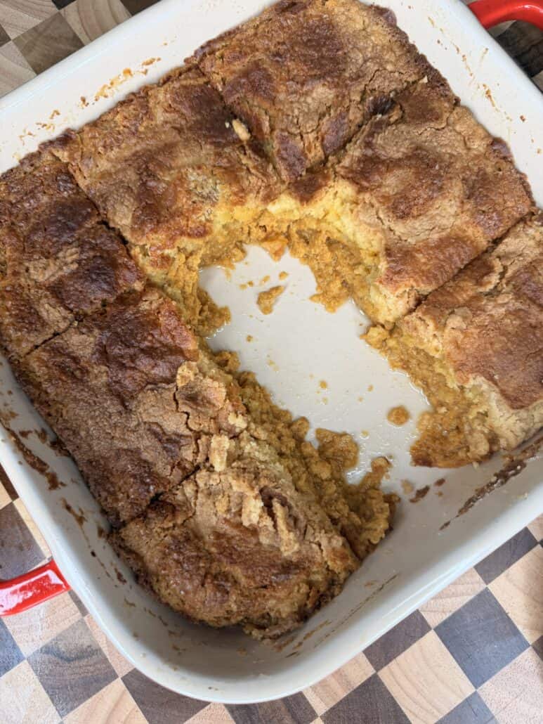 A partially eaten casserole dish filled with golden-brown, crumbly Pumpkin Pie Cake sits on a wooden surface, showing a missing section from one corner.
