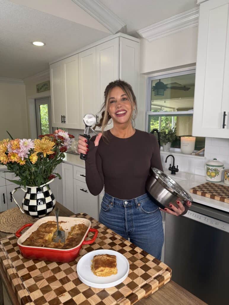 A smiling woman in a kitchen holds a metal bowl and ice cream scoop. She stands by a cutting board with a casserole dish, a plate topped with Pumpkin Pie Cake, and a vase of flowers.