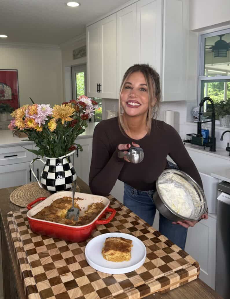 A smiling woman stands in a kitchen holding a bowl of whipped cream and an ice cream scoop next to a plate and a red dish of Pumpkin Pie Cake on the counter, with a vase of flowers nearby.