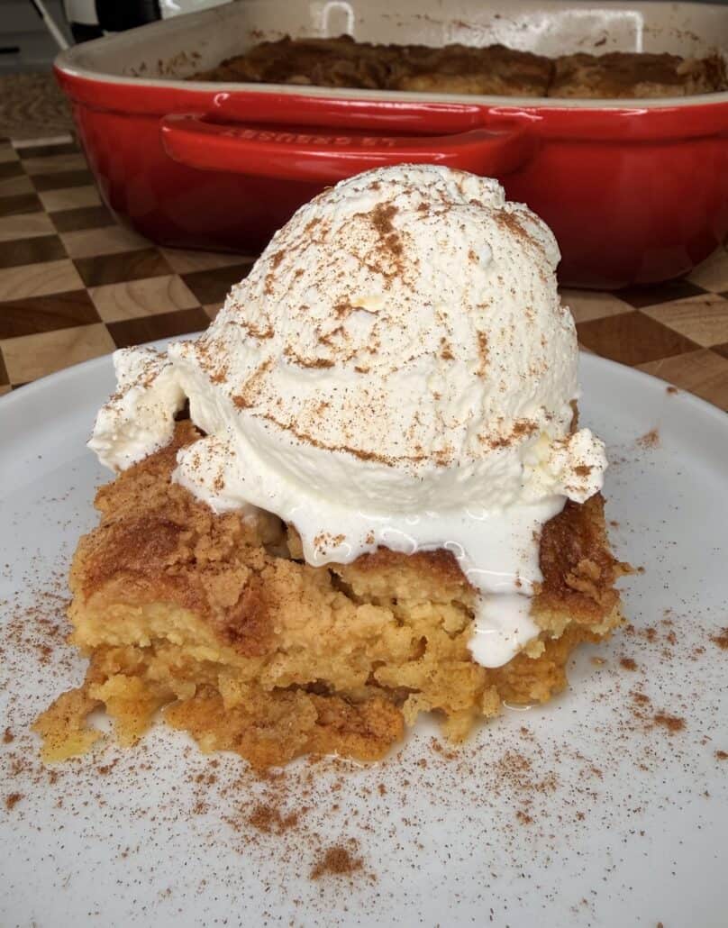 A slice of Pumpkin Pie Cake dump cake topped with a scoop of vanilla ice cream and a sprinkle of cinnamon, served on a white plate; a red baking dish is in the background.