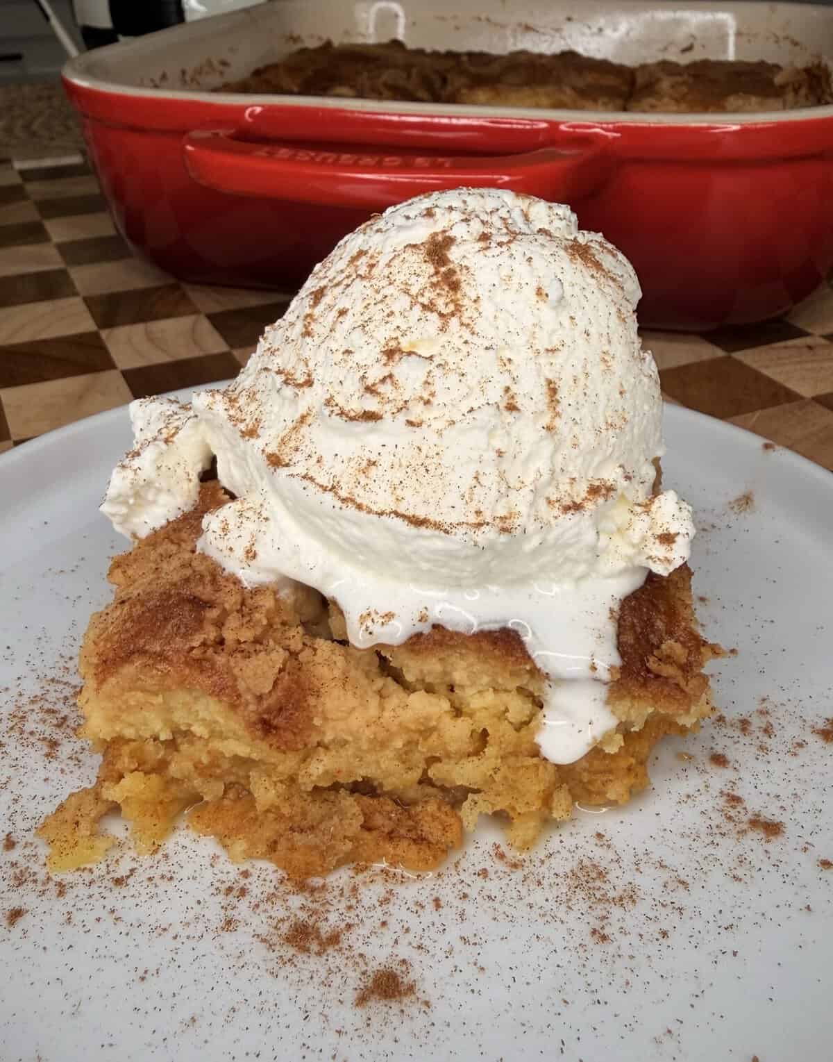 A slice of Pumpkin Pie Cake dump cake topped with a scoop of vanilla ice cream and a sprinkle of cinnamon, served on a white plate; a red baking dish is in the background.