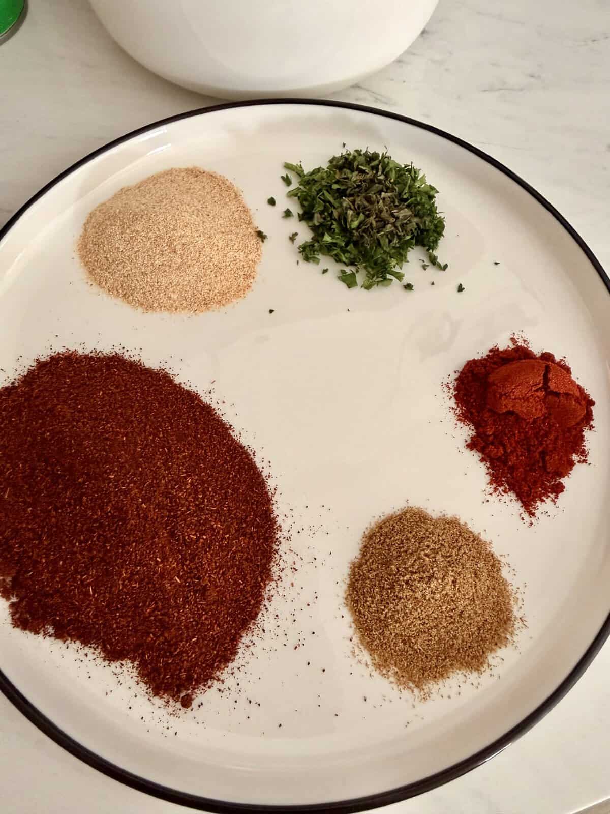 A plate with four piles of spices and herbs: a large pile of deep red chili powder, a pile of tan powder, a smaller pile of bright red powder, and a pile of chopped green herbs.