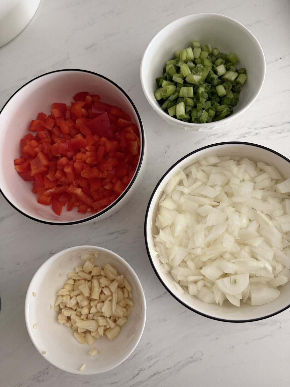 Four bowls on a marble countertop containing ingredients for chili: chopped red bell peppers, chopped white onions, sliced green onions, and minced garlic.