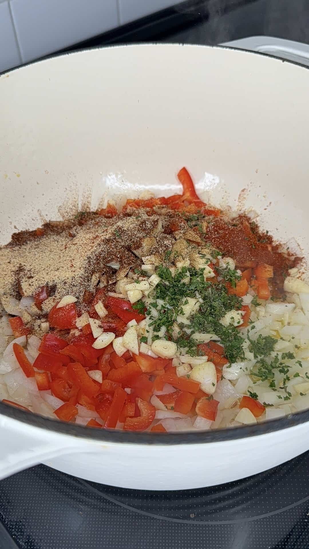 Chopped onions, bell peppers, garlic, and fresh herbs topped with various chili spices in a white enameled pot.