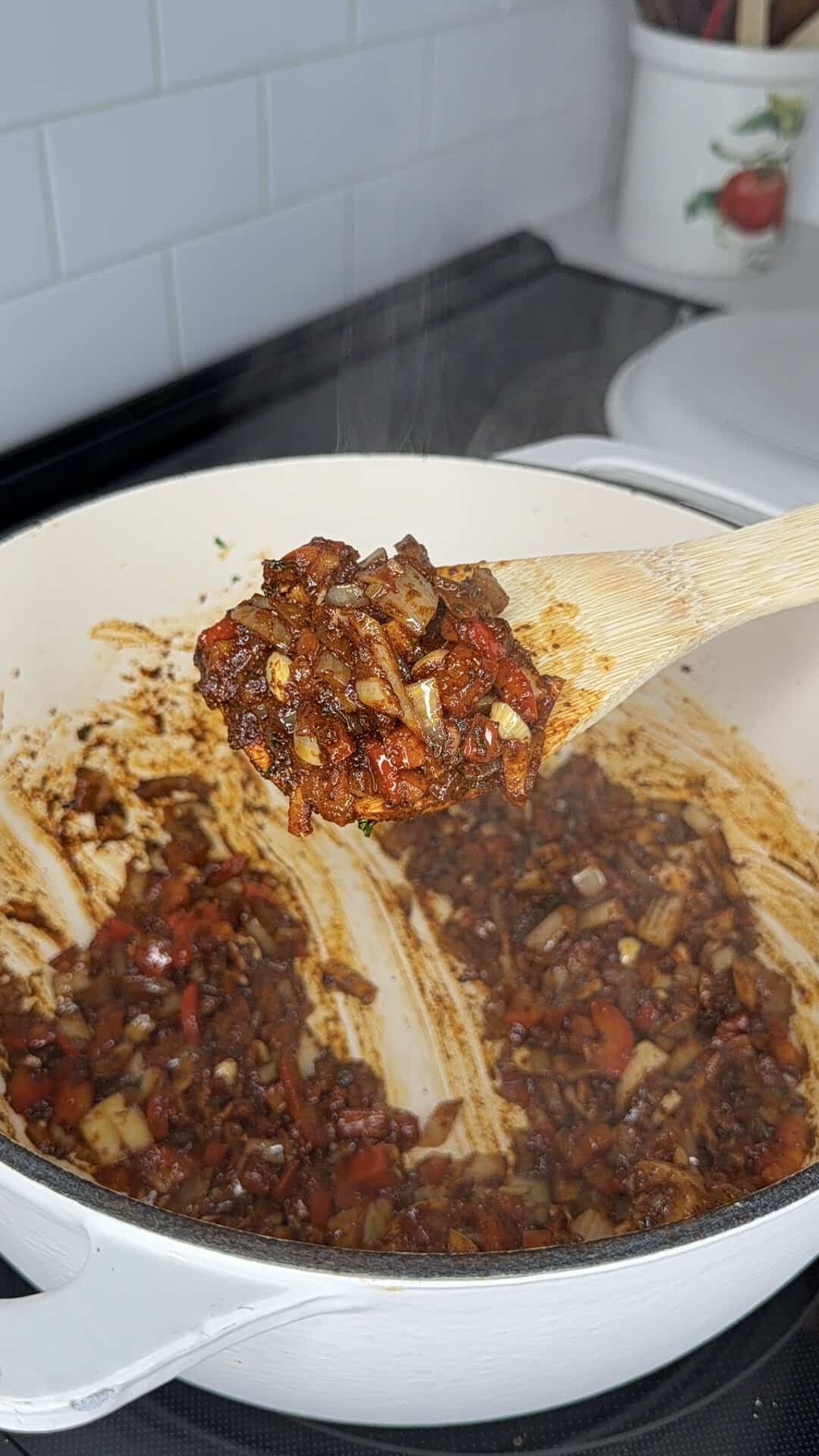A wooden spoon holds a mixture of sautéed onions and diced red peppers for chili over a large white cooking pot containing more of the same mixture.