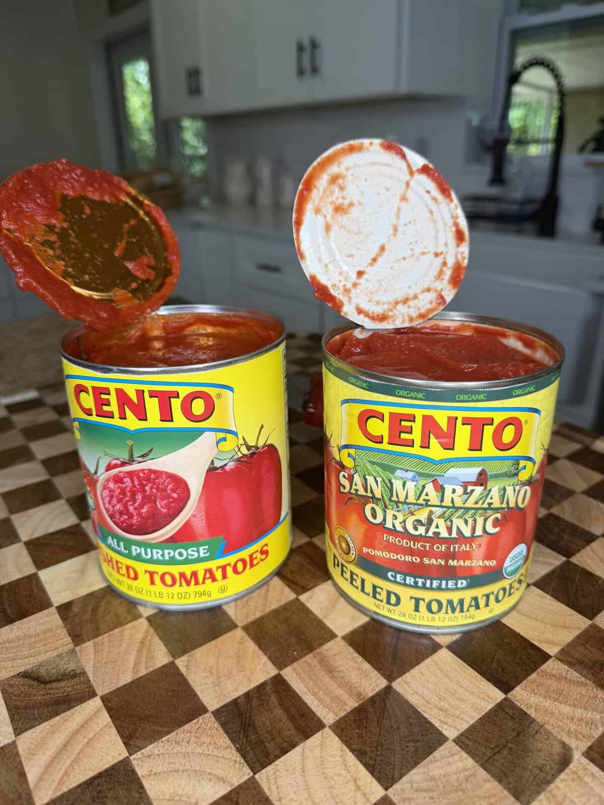 Two open cans of Cento tomatoes, one labeled crushed tomatoes and the other organic San Marzano peeled tomatoes, sitting on a checkered wooden surface ready for chili.
