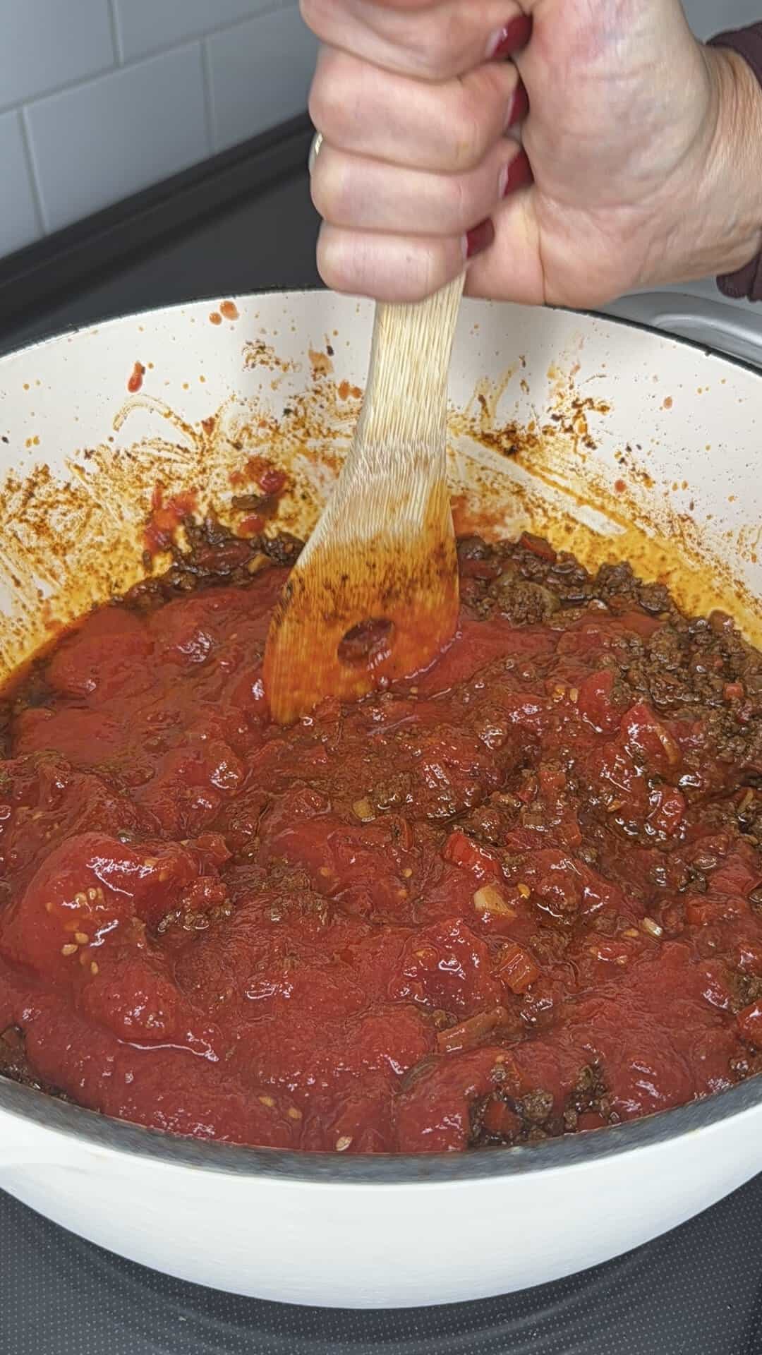 A hand holds a wooden spoon to stir chili made with tomato sauce and ground meat in a large pot.