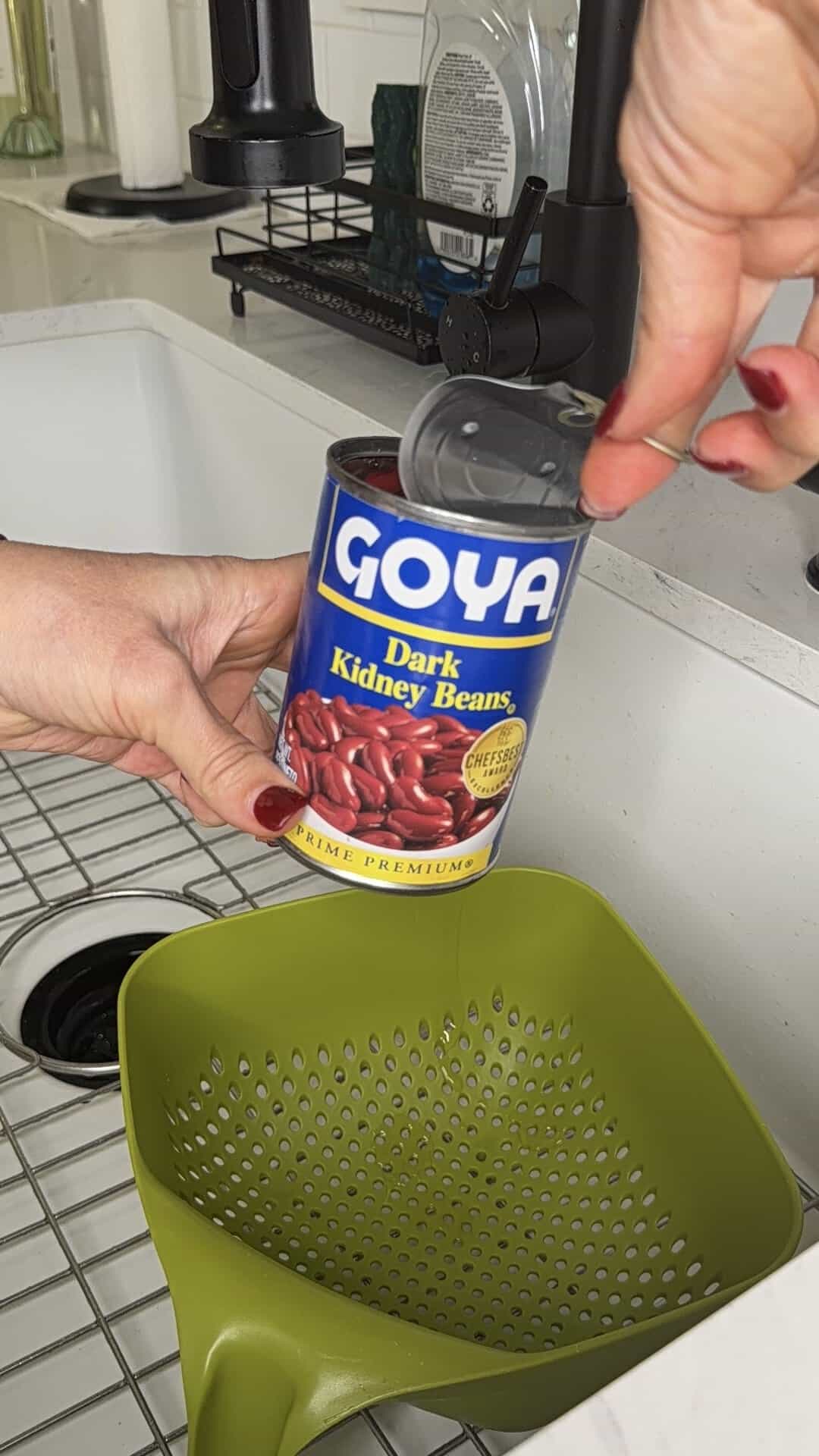 A person opens a can of Goya dark kidney beans over a green colander in a kitchen sink, preparing ingredients for chili.
