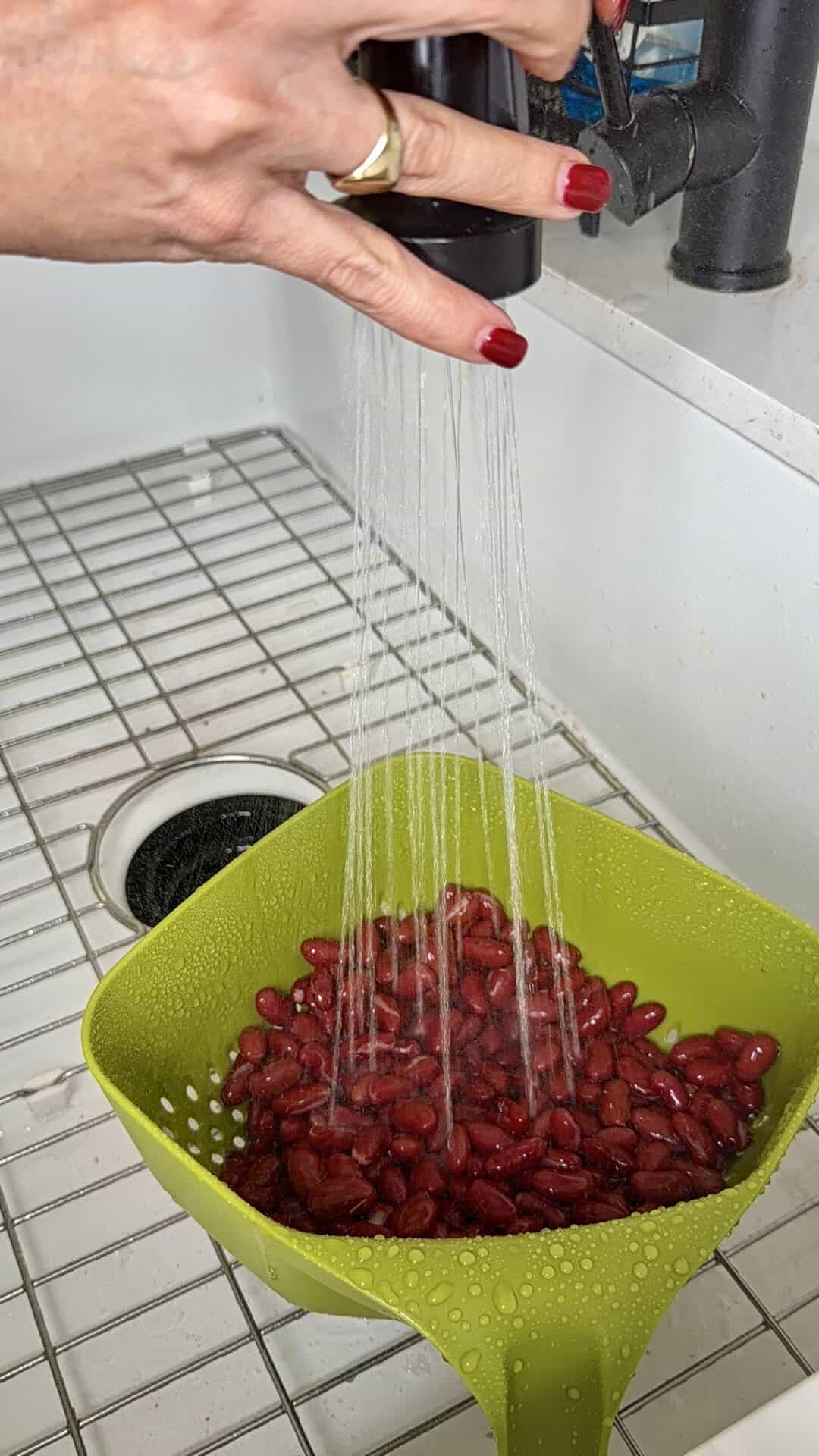 A hand rinsing red kidney beans for chili in a green colander under a kitchen faucet.