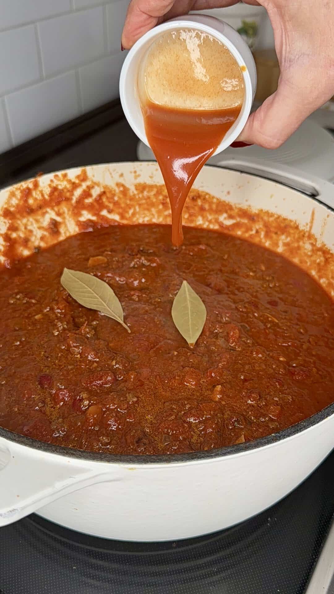 A hand pours red sauce from a small white cup into a white pot of simmering chili topped with two bay leaves.