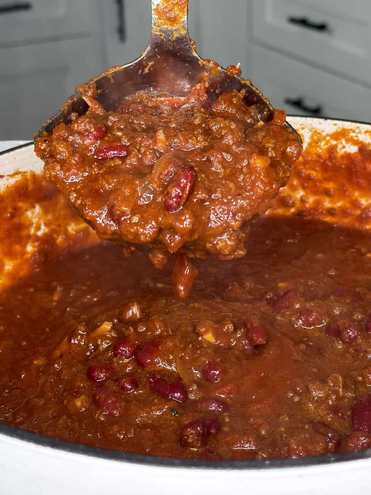 A ladle holds a portion of thick, red chili with beans above a large pot of the same dish.