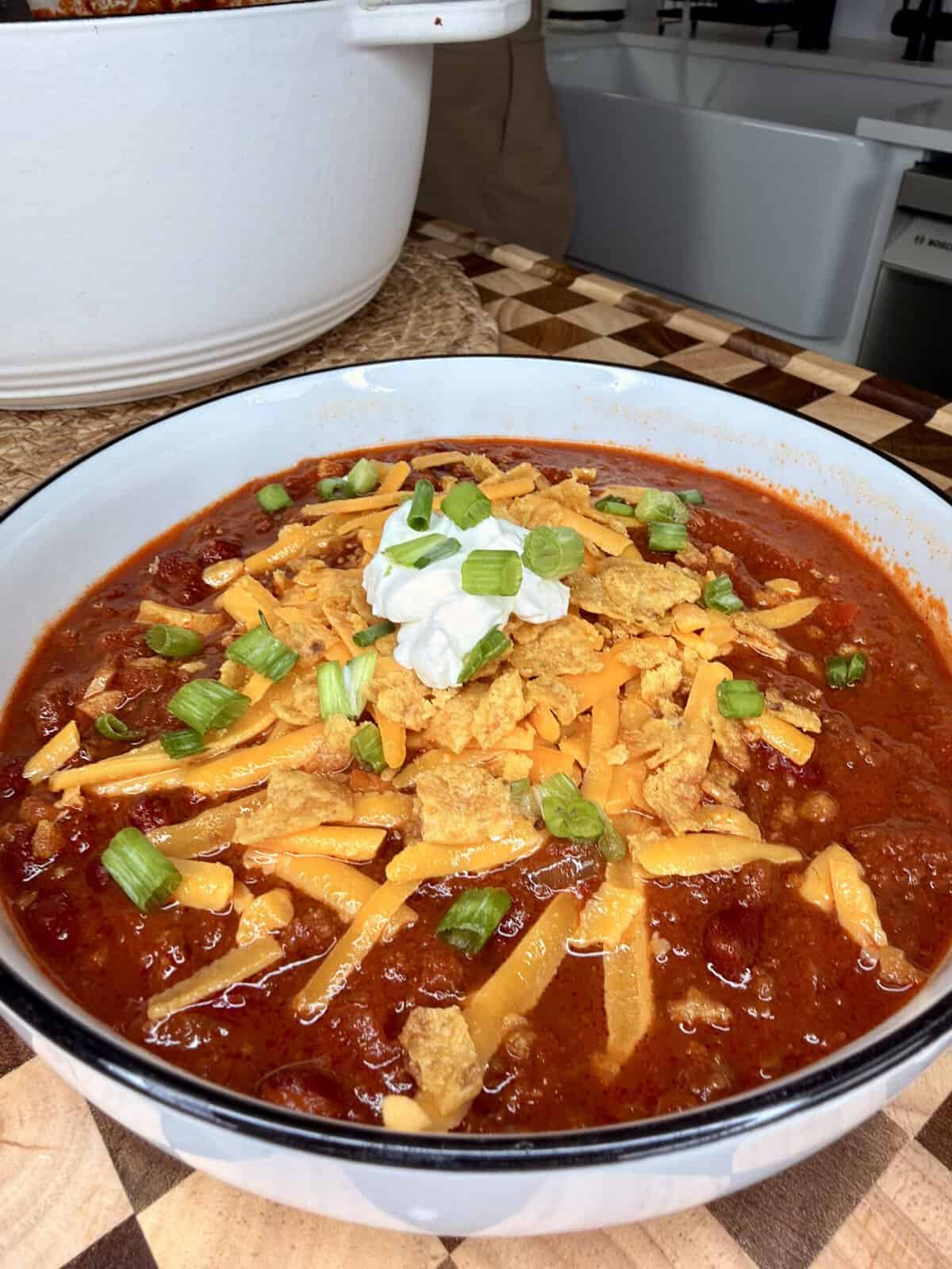 A bowl of chili topped with shredded cheddar cheese, crushed corn chips, a dollop of sour cream, and chopped green onions.