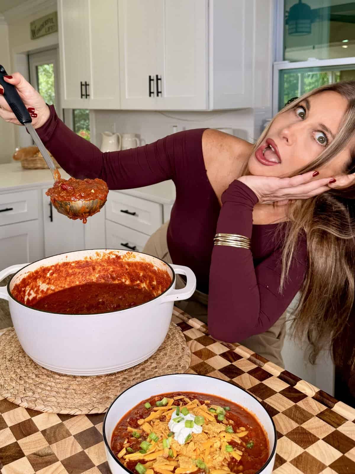 A woman holds a ladle of chili over a large white pot, with a bowl of chili topped with cheese, sour cream, and green onions in the foreground.