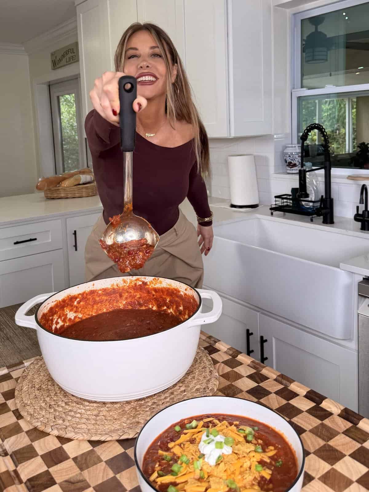 A woman in a kitchen holds a ladle filled with chili over a large white pot, with a bowl of chili topped with cheese, sour cream, and green onions in the foreground.