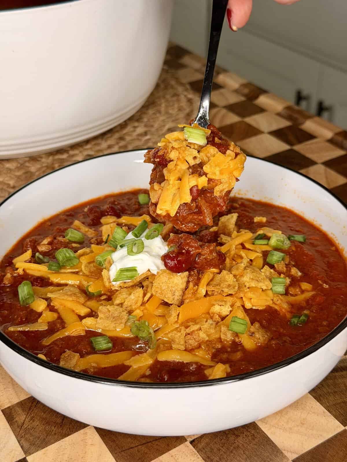 A spoonful of chili topped with shredded cheese, sour cream, and green onions held over a bowl of the same.