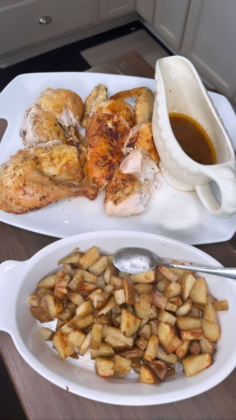 A platter of Whole Roasted Chicken pieces with a gravy boat of brown sauce sits behind golden, diced roasted potatoes. Both dishes rest on a wooden table in a cozy kitchen setting.