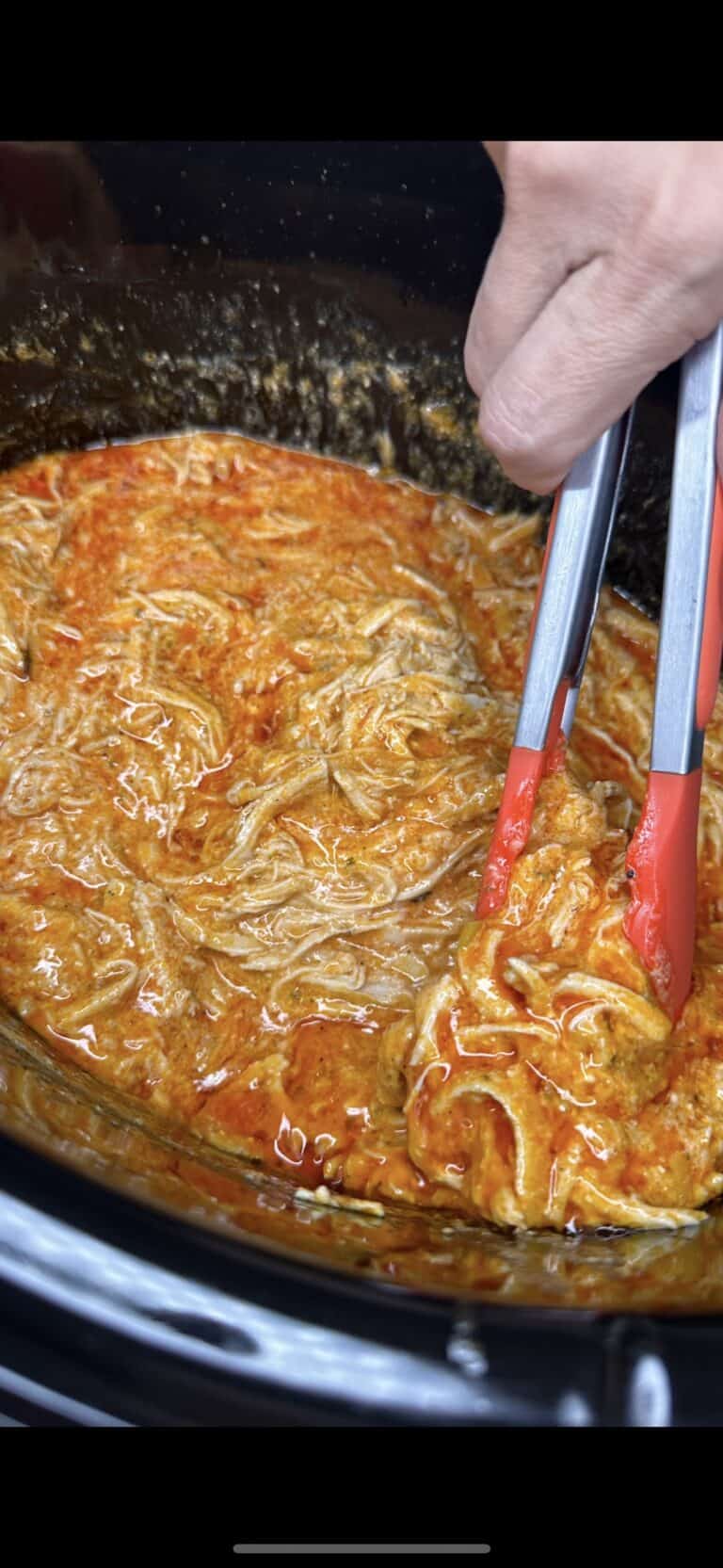 A close-up of Crockpot Buffalo Chicken Dinner shows shredded meat in a reddish-orange sauce inside a slow cooker, with a hand using red-handled tongs to lift some tender chicken.