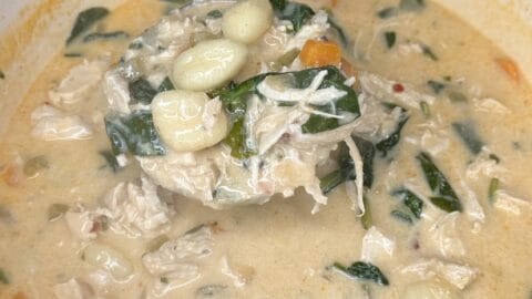 A ladle lifts a serving of creamy Chicken and Gnocchi Soup with spinach, carrots, and shredded chicken from a pot on a kitchen counter.
