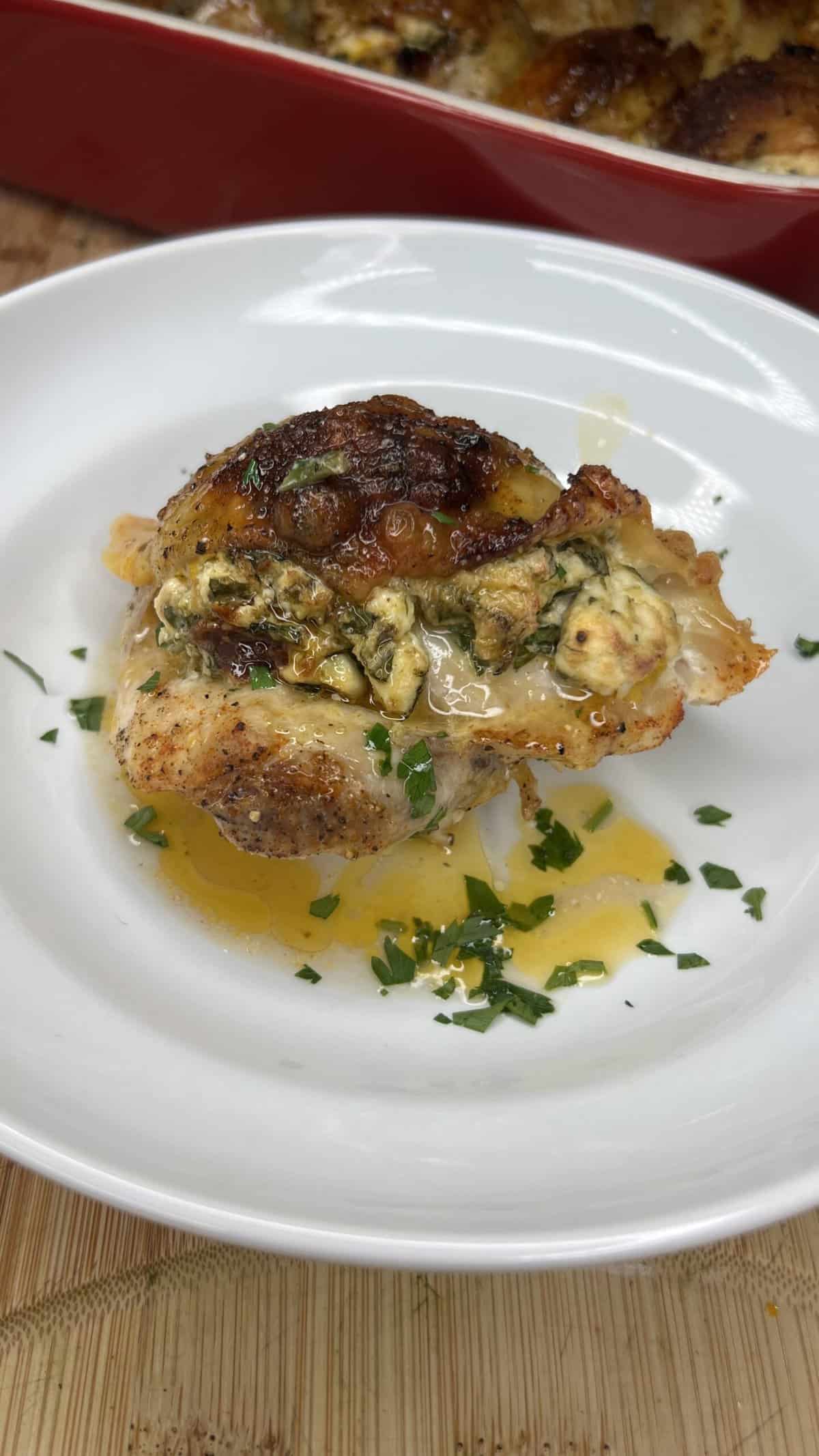 A cooked, stuffed chicken thigh sits on a white plate, topped with melted cheese and herbs, with juices pooling underneath and a sprinkle of chopped parsley for garnish.