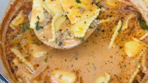A ladle lifts cheesy ravioli, sausage, and herbs from a creamy tomato soup in a white pot. This One Pot Sausage Ravioli Soup is rich and orange-red, with visible spices and chopped greens for extra flavor.