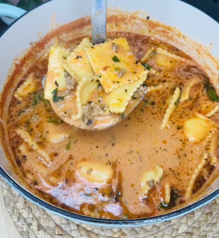 A ladle lifts cheesy ravioli, sausage, and herbs from a creamy tomato soup in a white pot. This One Pot Sausage Ravioli Soup is rich and orange-red, with visible spices and chopped greens for extra flavor.