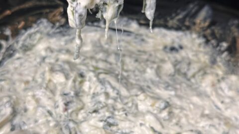 A close-up of a spoon lifting creamy crockpot spinach artichoke dip from a slow cooker, with a bag of tortilla chips visible in the background.