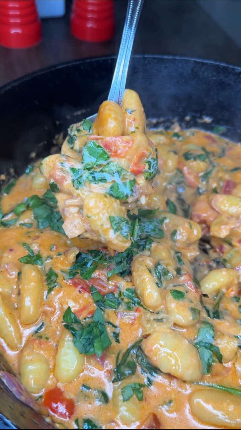 Close-up of creamy gnocchi in a skillet, garnished with chopped greens. A spoonful of gnocchi, coated in Melissa Jo's Signature Cream Sauce with herbs and vegetables, is being lifted from the pan.