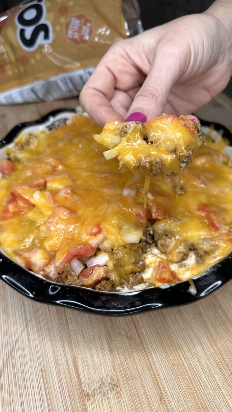 A hand holding a cheesy serving of Turkey Taco Dip casserole above a dish filled with layers of melted cheese, ground turkey, tomatoes, and other ingredients, with a bag of tortilla chips in the background.