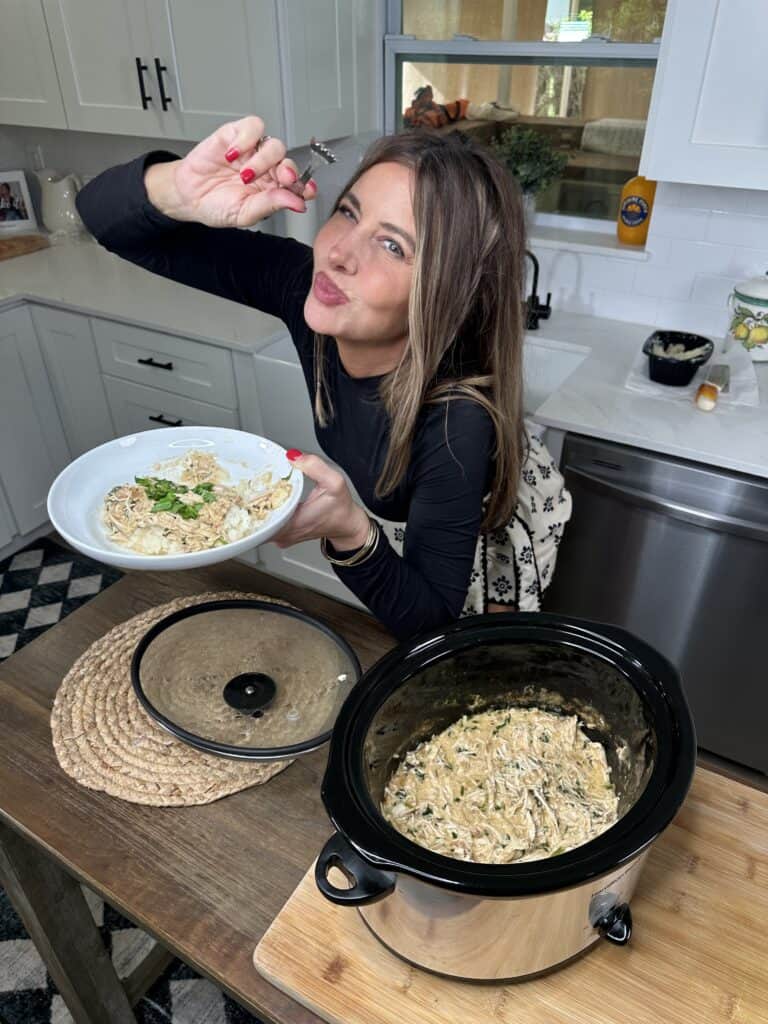 A woman in a kitchen holds a plate of creamy pasta and playfully pretends to eat with her fingers, standing beside a slow cooker filled with Creamy Dijon Crockpot Chicken. She looks enthusiastic and happy.