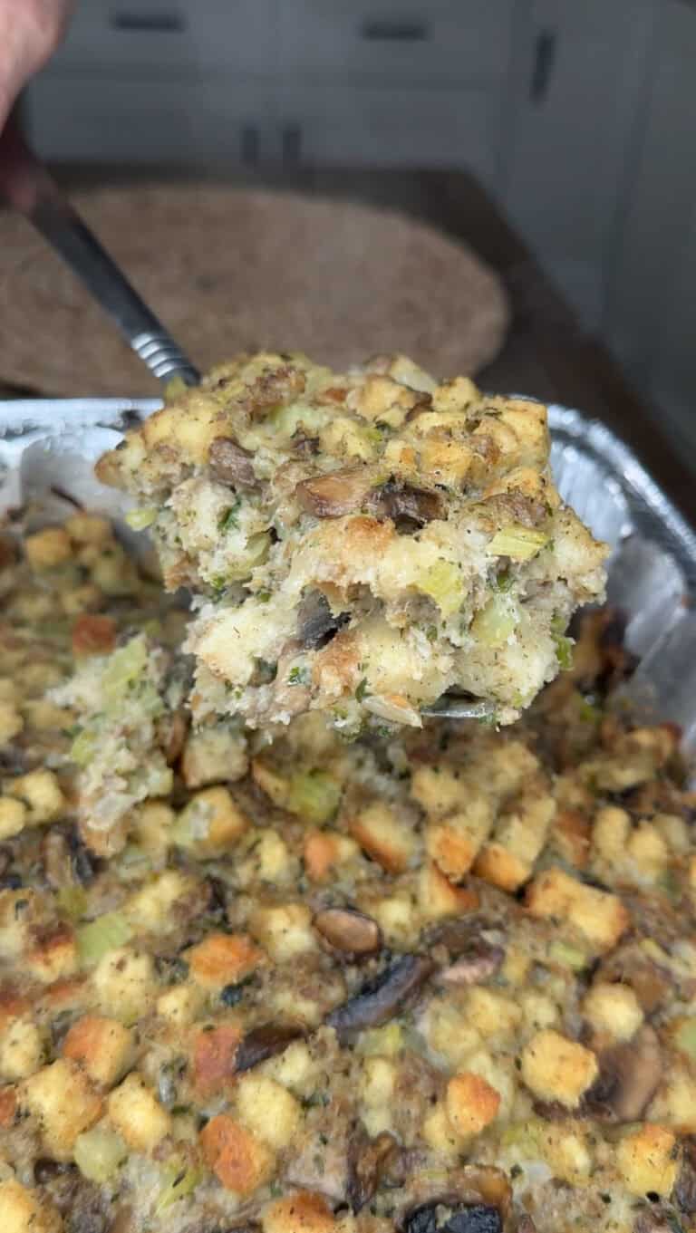 A close-up of Traditional Sausage Stuffing with herbs, celery, and mushrooms being scooped from a foil baking pan. The stuffing appears moist and flavorful, with visible chunks of bread, sausage, and vegetables.