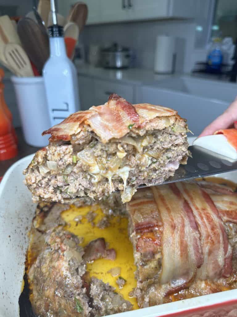 A slice of The Best Meatloaf Ever—bacon-wrapped with melted cheese inside—is being lifted from a white baking dish on a kitchen counter, surrounded by utensils and appliances in the background.
