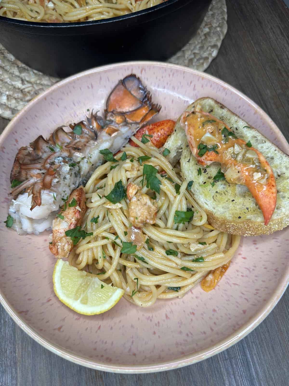 A plate of Lobster Pasta with lobster pieces, garnished with parsley, served alongside half a lobster tail, a slice of garlic bread, and two lemon wedges on a pink dish atop a wooden table.