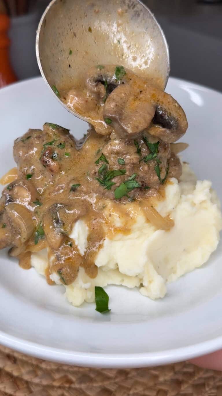 A spoon pours creamy mushroom and herb sauce over tender Meatball Stroganoff, served atop a mound of mashed potatoes in a white bowl.