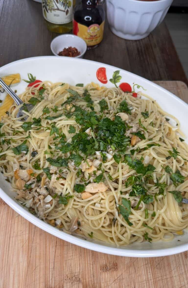 A large oval dish filled with linguine and clams, mixed with chopped herbs and garnished with fresh parsley, sits on a wooden surface. A fork rests in the dish, with condiments and a bowl in the background.