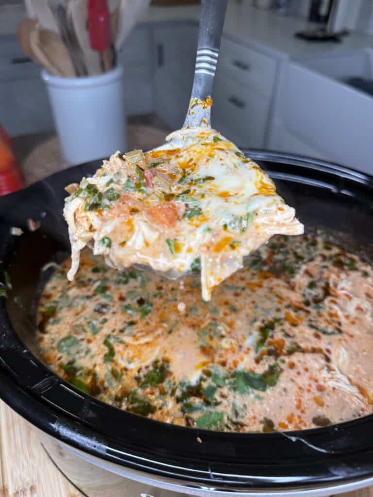 A close-up of a ladle lifting a cheesy, Chicken Parmesan Soup-inspired, tomato-based lasagna soup with pasta, spinach, and herbs from a black slow cooker in a cozy kitchen setting.