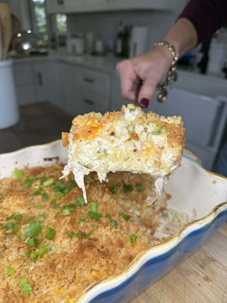 A hand lifts a serving of Heavenly Chicken and Rice Casserole with a golden crumb topping, revealing creamy filling with bits of vegetables. The casserole is garnished with chopped green onions in a cozy kitchen setting.