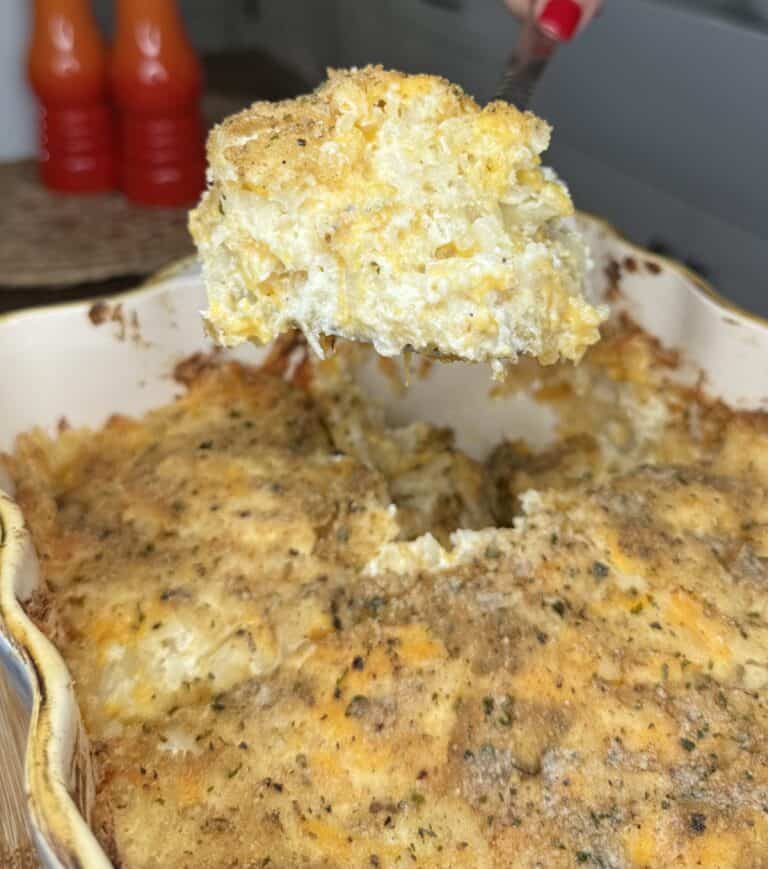Close-up of a baked macaroni and cheese casserole with a golden, crispy breadcrumb topping. A serving is being lifted out, revealing creamy macaroni with melted cheese—perfect comfort food alongside Party Potatoes. Two orange bottles are blurred in the background.