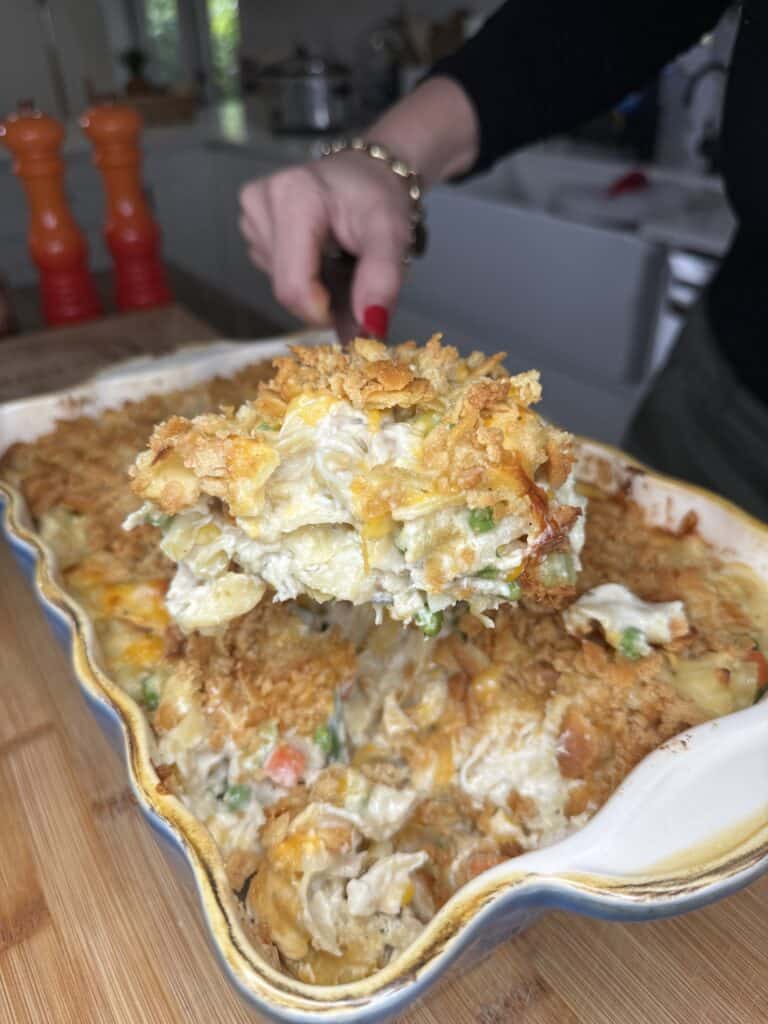 A hand serves a large, cheesy portion of Creamy Chicken Noodle Casserole from a baking dish filled with vegetables and topped with a golden, crunchy crust. The background shows a cozy kitchen scene.