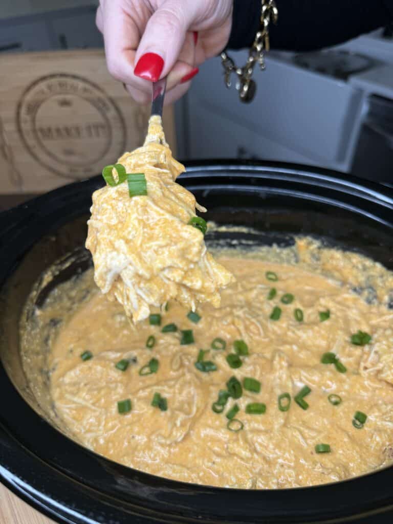 A hand with red nail polish lifts a forkful of creamy Crockpot Buffalo Chicken Dip from a slow cooker, garnished with chopped green onions.