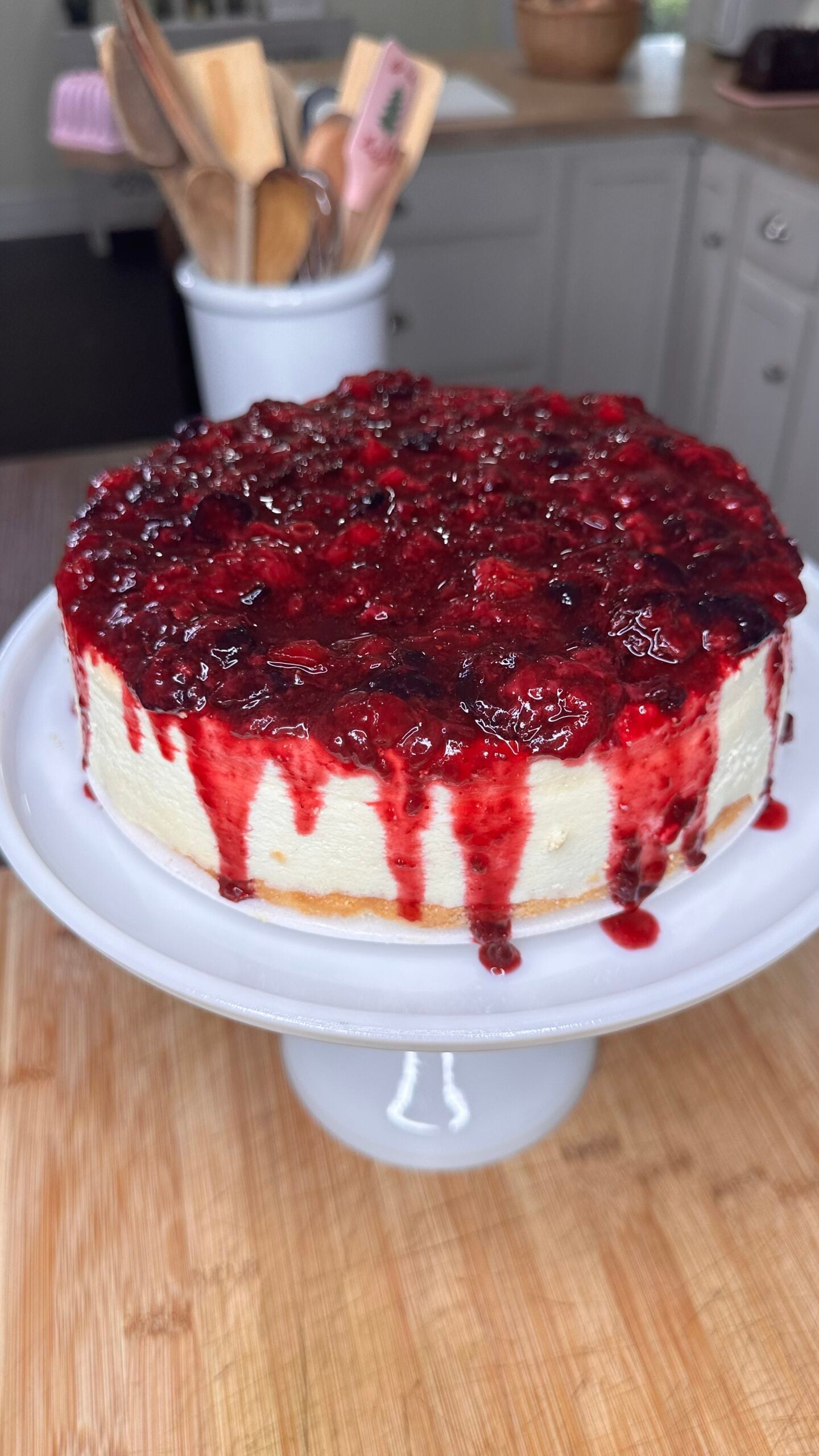 A creamy cheesecake topped with Melissa Jo's Berry Compote, a thick, glossy mixed berry sauce that drips down the sides, displayed on a white cake stand in a kitchen setting.