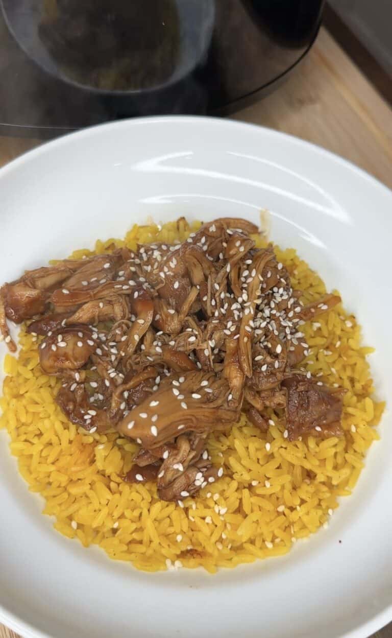 A white bowl filled with yellow rice, topped with Crockpot Honey Garlic Chicken in a savory brown sauce and sprinkled with white sesame seeds.