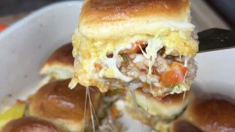 A close-up of a Sausage and Peppers Sliders breakfast sandwich with melted cheese, sausage, egg, and peppers in a soft roll, held above a baking dish filled with more delicious sliders.