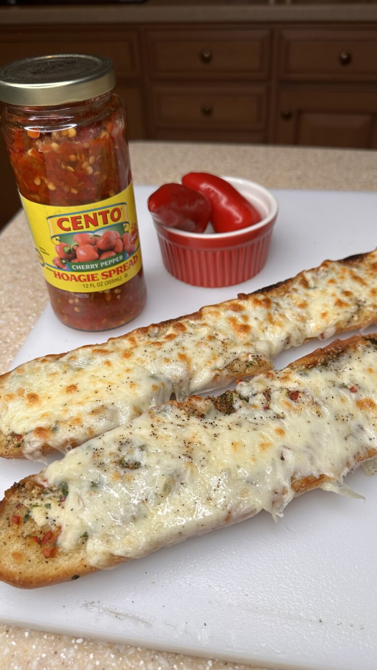 Two pieces of Cherry Pepper Garlic Bread sit on a white cutting board, with a jar of Cento Cherry Pepper Hoagie Spread and a small bowl of fresh red peppers in the background.