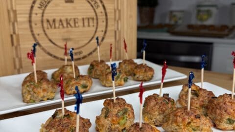 Two white plates hold savory Party Balls, each pierced with decorative toothpicks. In the background, a wooden cutting board engraved with “Make It!” stands upright in a modern kitchen setting.