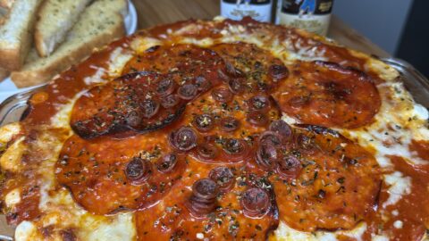 A delicious Pepperoni Pizza Dip, baked with cheesy goodness and topped with pepperoni slices and herbs, sits on a wooden surface alongside garlic bread and two Dan-O’s seasoning bottles in the background.