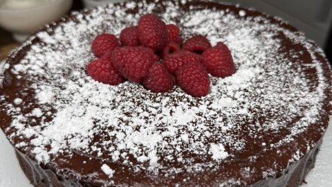 A Flourless Chocolate Torte topped with fresh raspberries and dusted with powdered sugar sits on a white plate in a modern kitchen.