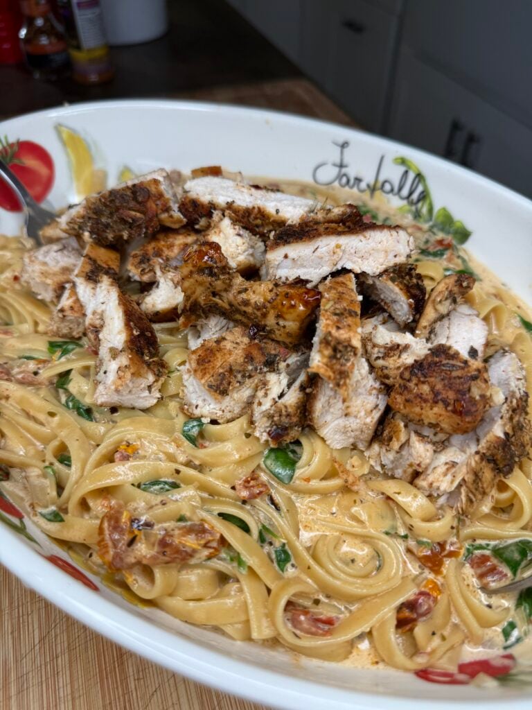 A close-up of Chicken Creamy Fettuccine with sun-dried tomatoes, spinach, and sliced grilled chicken breast on top, served in a decorative bowl on a kitchen counter.