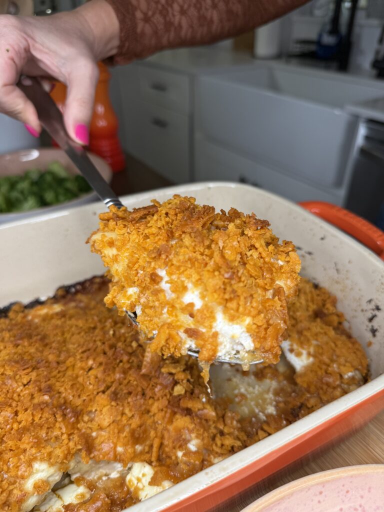 A hand lifts a serving of Cheez-It Chicken casserole, topped with a crunchy orange layer, from a rectangular baking dish in the kitchen.