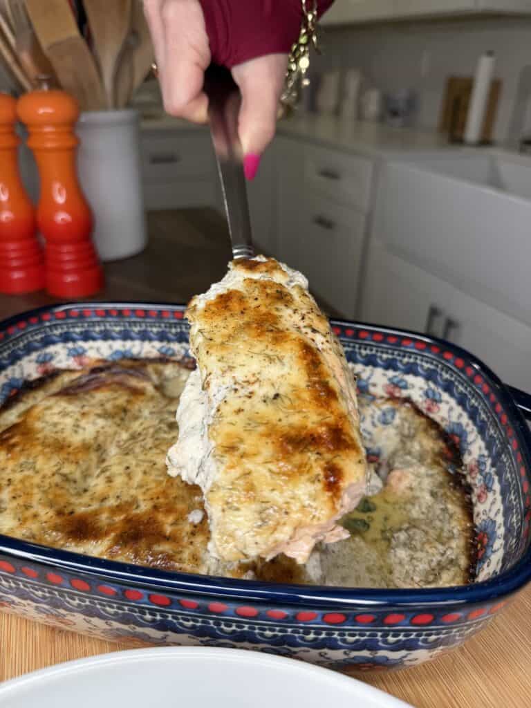 A hand holding a spatula lifts a piece of Dilly Salmon, covered in creamy, browned sauce, from a blue patterned baking dish in a kitchen setting.