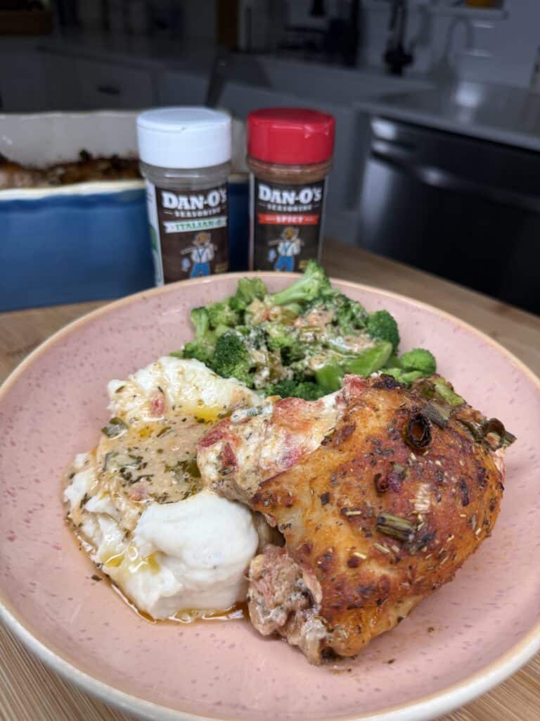 A plate of Fire Roasted Stuffed Chicken Thighs, mashed potatoes with gravy, and steamed broccoli sits on a pink dish. Two Dan-O’s seasoning bottles and a casserole dish are visible in the background.